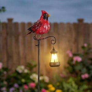 May include: A decorative garden stake featuring a red cardinal bird perched atop a black metal pole. The pole has a decorative lantern with a warm, inviting glow. The stake is surrounded by colorful flowers and greenery.