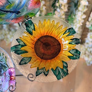 May include: A clear glass decorative plate with a sunflower design, featuring yellow petals and a brown centre. Green leaves surround the sunflower. The plate has a textured surface and is supported by a black metal stand. Other plates with floral designs are also visible.