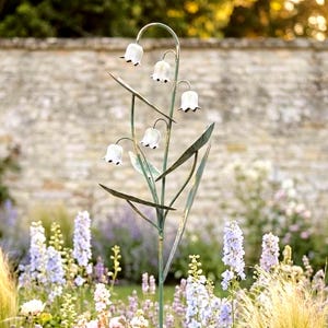 May include: A metal garden stake shaped like a lily of the valley plant. The stake has a green stem with leaves and white bell-shaped flowers with dark accents. The stake is in a garden with various flowers.