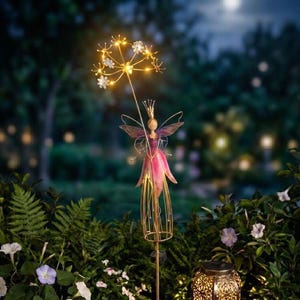 May include: A whimsical garden stake featuring a fairy with a pink dress and a crown, holding a glowing flower. A solar-powered lantern with a decorative pattern is also visible, surrounded by lush greenery and flowers, creating a magical ambiance.
