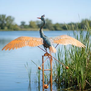 A metal sculpture of a heron stands in the water, its wings spread wide, with tall reeds and trees in the background.