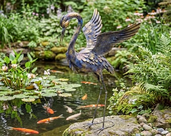 May include: A metal garden sculpture of a heron with outstretched wings, featuring a weathered blue and bronze finish with textured feather details. The bird stands on thin metal legs, positioned on a mossy rock beside a pond with koi fish.