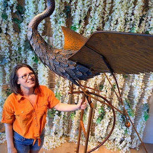 A woman in an orange shirt stands next to a large metal sculpture of a bird, which appears to be a large metal bird with a long neck and legs.