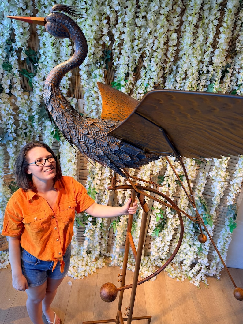 A woman in an orange shirt stands next to a large metal sculpture of a bird, which appears to be a large metal bird with a long neck and legs.
