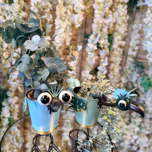 May include: Two light blue metal planters with bird-like faces, each holding greenery. The planters have gold rims and are mounted on black metal stands. The background features white floral arrangements.