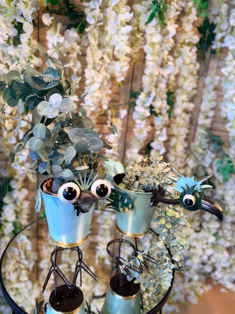 May include: Two light blue metal planters with bird-like faces, each holding greenery. The planters have gold rims and are mounted on black metal stands. The background features white floral arrangements.