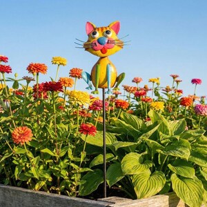 A colorful metal cat figurine is perched on a metal pole in a garden filled with vibrant flowers.