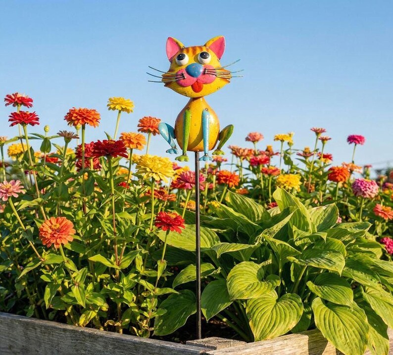 A colorful metal cat figurine is perched on a metal pole in a garden filled with vibrant flowers.