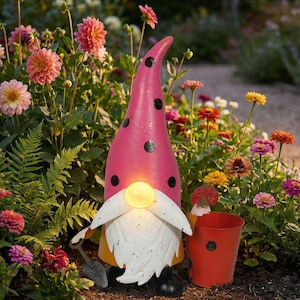 Könnte beinhalten: Eine skurrile Gartenzwerg-Statue mit einem rosa Hut mit schwarzen Tupfen. Der Zwerg hat einen weißen Bart, ein gelbes Licht für die Nase und hält einen kleinen roten Eimer. Die Statue ist von bunten Blumen umgeben.