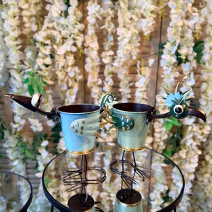 May include: Two metal bird planters with light blue and green buckets, perched on black stands. The birds have detailed metal wings, beaks, and eyes. The background features white flowers.