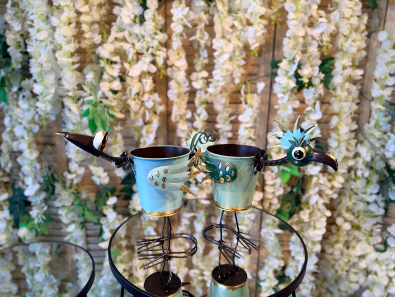 May include: Two metal bird planters with light blue and green buckets, perched on black stands. The birds have detailed metal wings, beaks, and eyes. The background features white flowers.