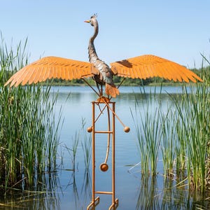 A large metal sculpture of a crane-like bird with orange wings stands on a metal stand in a body of water, surrounded by tall reeds.