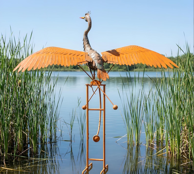 A large metal sculpture of a crane-like bird with orange wings stands on a metal stand in a body of water, surrounded by tall reeds.