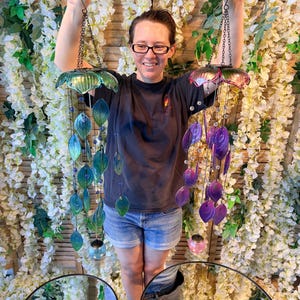 A woman is standing in front of a floral backdrop, holding up two colorful wind chimes with green and purple shades.