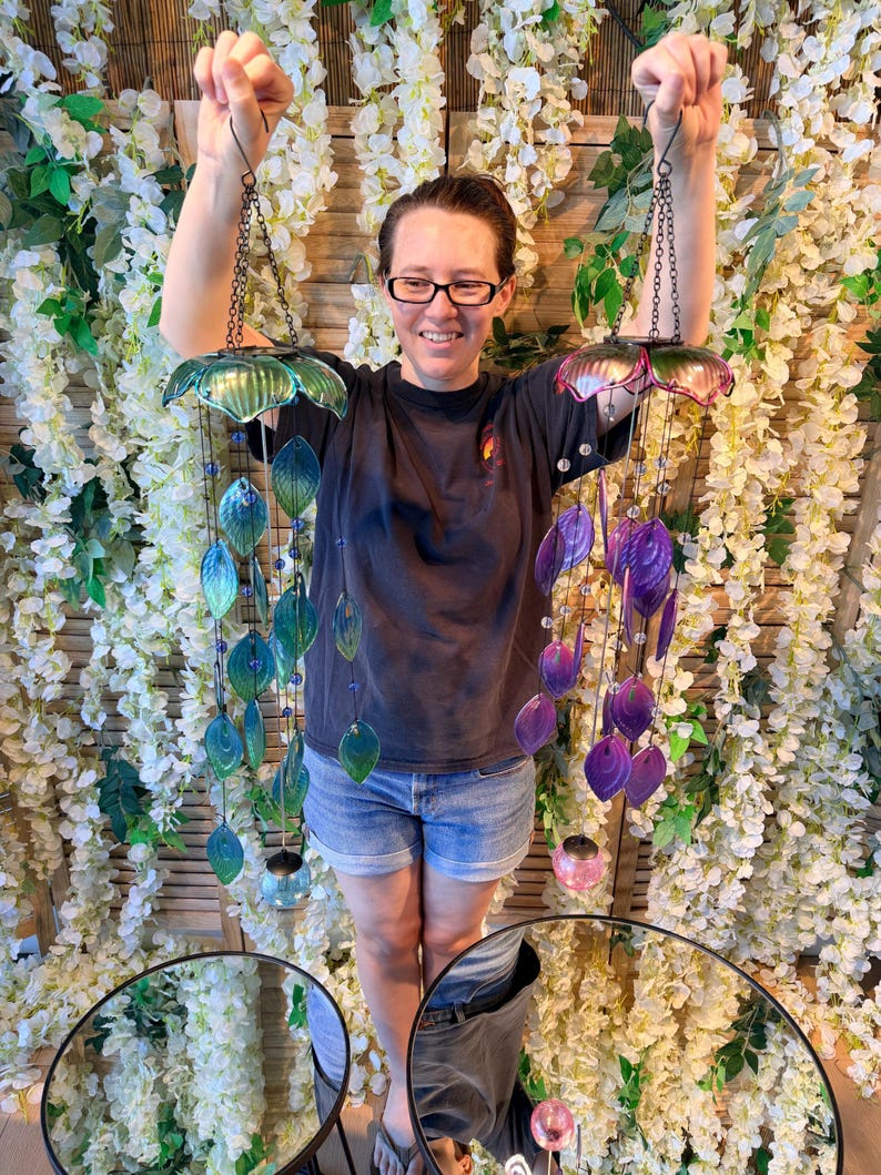 A woman is standing in front of a floral backdrop, holding up two colorful wind chimes with green and purple shades.