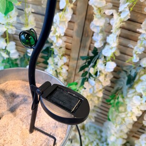 A solar-powered lamp with a green glass shade is placed on a wooden surface, surrounded by white flowers and greenery.