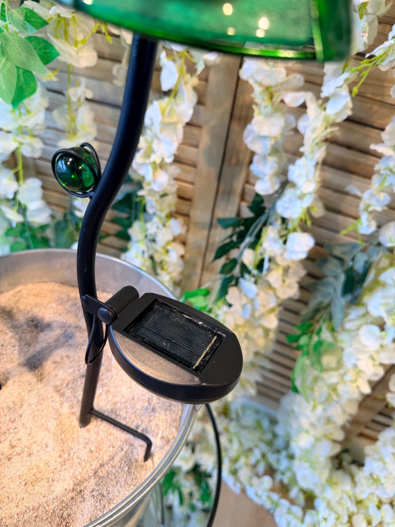 A solar-powered lamp with a green glass shade is placed on a wooden surface, surrounded by white flowers and greenery.