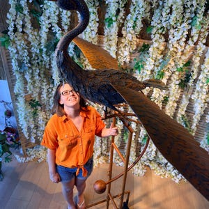 A woman in an orange shirt stands next to a large metal sculpture of a bird, which appears to be a windmill-like structure.
