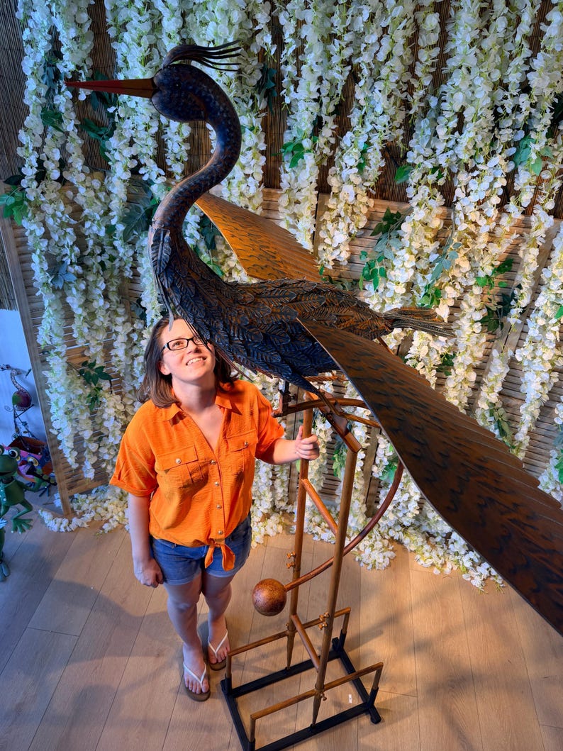 A woman in an orange shirt stands next to a large metal sculpture of a bird, which appears to be a windmill-like structure.