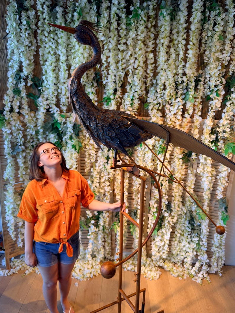 A woman in an orange shirt stands next to a large metal sculpture of a bird, which appears to be a decorative piece.