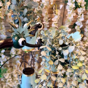 May include: A whimsical, light blue metal dragon planter with large googly eyes and gold accents, holding a small pot of trailing greenery. The planter sits on a mirrored surface, surrounded by white floral decor.