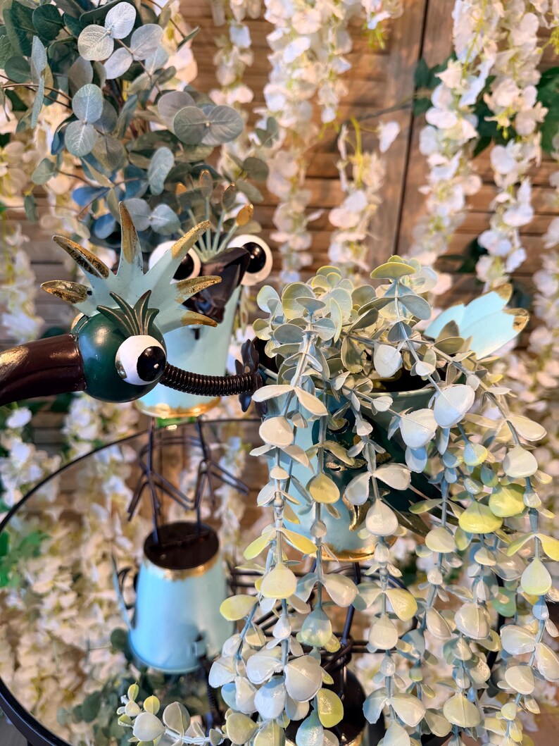 May include: A whimsical, light blue metal dragon planter with large googly eyes and gold accents, holding a small pot of trailing greenery. The planter sits on a mirrored surface, surrounded by white floral decor.