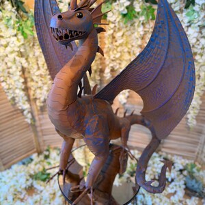 A metal dragon statue with blue wings and a brown body stands on a metal stand, surrounded by white flowers.