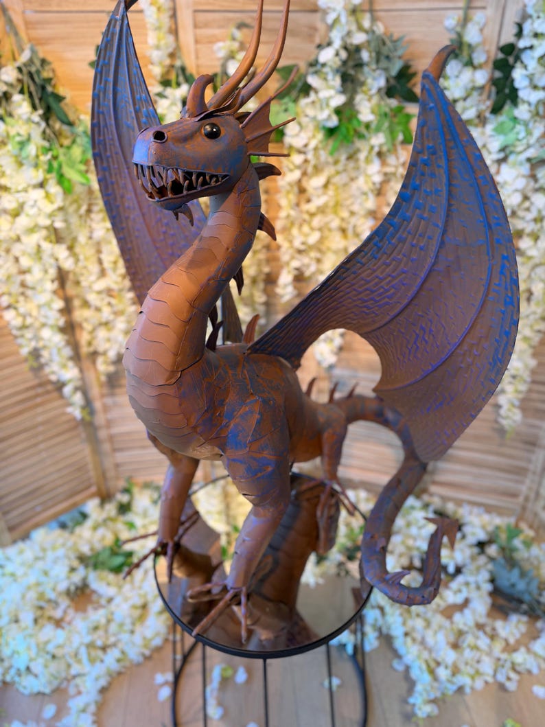 A metal dragon statue with blue wings and a brown body stands on a metal stand, surrounded by white flowers.