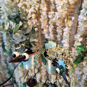 May include: Two whimsical bird-shaped planters with googly eyes and long beaks. The light blue metal planters hold green leafy plants. The planters are on a round, mirrored surface, with a backdrop of white flowers.