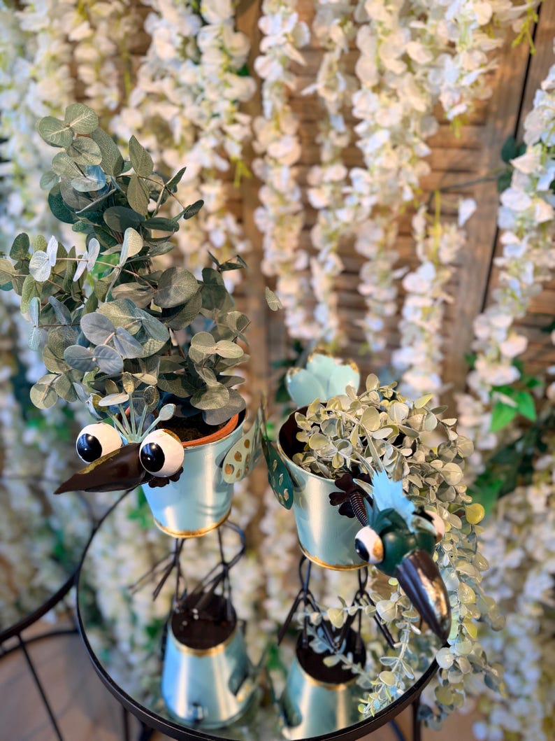 May include: Two whimsical bird-shaped planters with googly eyes and long beaks. The light blue metal planters hold green leafy plants. The planters are on a round, mirrored surface, with a backdrop of white flowers.