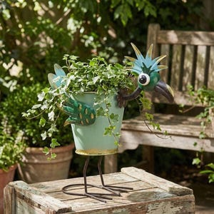 A metal bird sculpture with a green body and blue head is perched on a wooden crate, holding a plant in a pot.