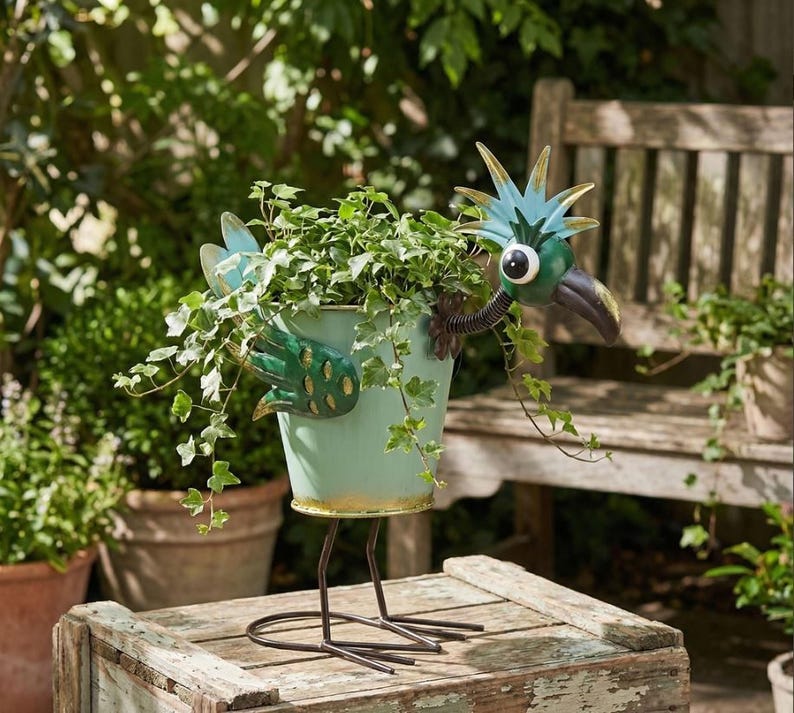 A metal bird sculpture with a green body and blue head is perched on a wooden crate, holding a plant in a pot.