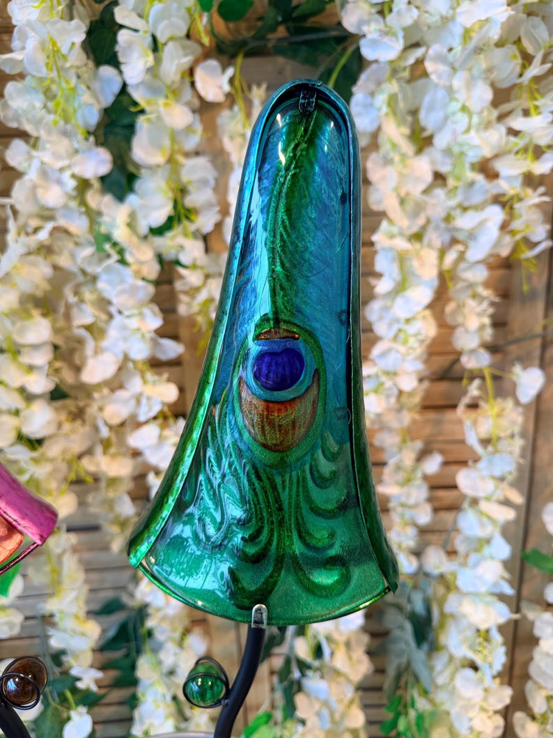 A colorful glass sculpture with a peacock feather design, set against a backdrop of white flowers.