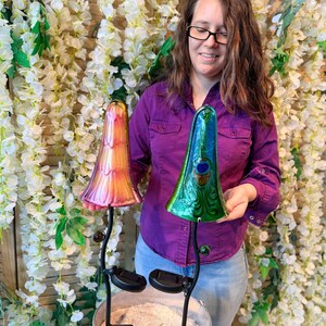 A woman in a purple shirt is holding two colorful glass mushroom-shaped lamps, one pink and one green, in front of a backdrop of white flowers.