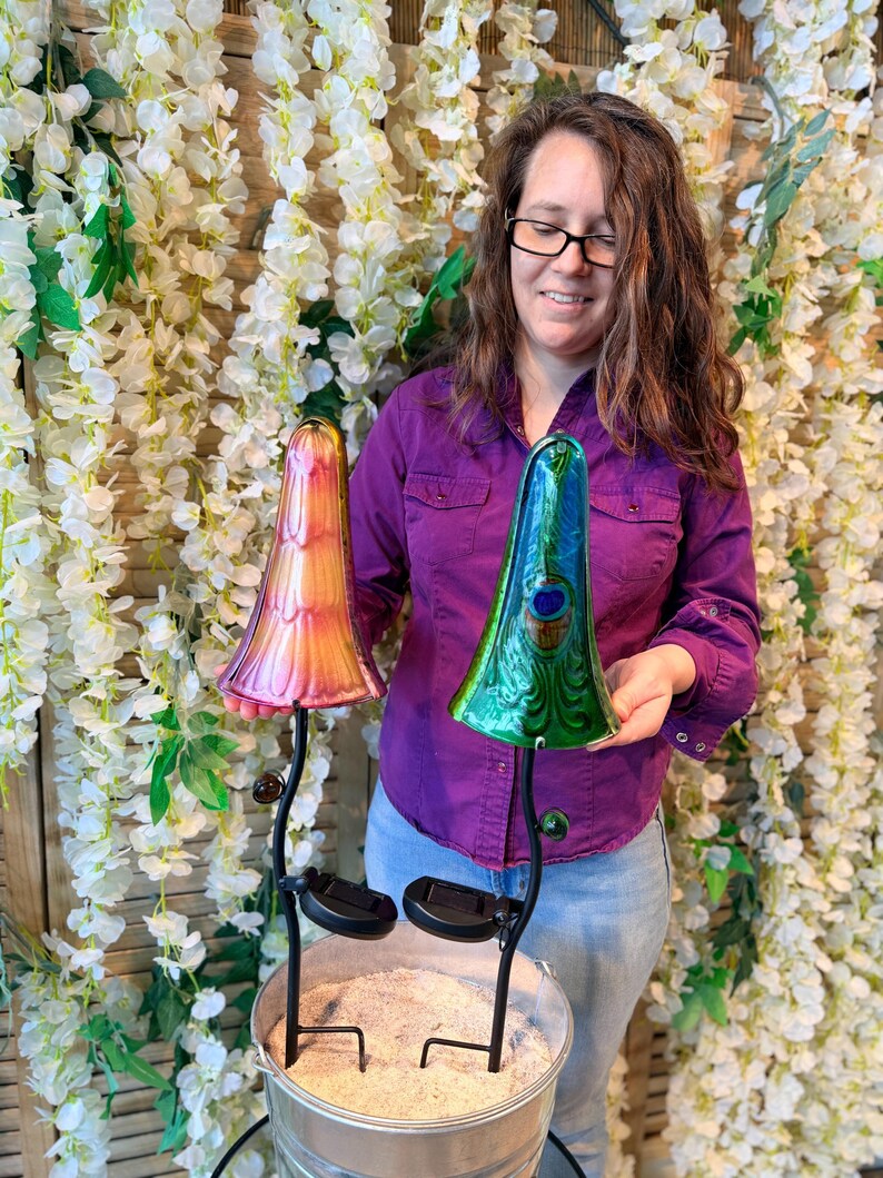 A woman in a purple shirt is holding two colorful glass mushroom-shaped lamps, one pink and one green, in front of a backdrop of white flowers.