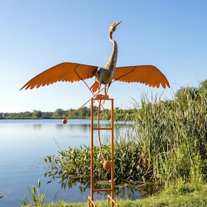 A large metal sculpture of a bird with orange wings and a long neck stands on a metal stand near a body of water, surrounded by tall grass.