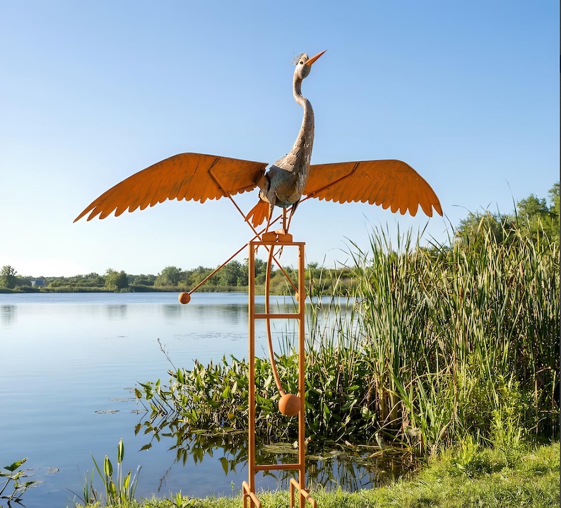 A large metal sculpture of a bird with orange wings and a long neck stands on a metal stand near a body of water, surrounded by tall grass.