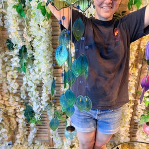 A woman is smiling and holding a colorful glass wind chime with green and blue leaves hanging from it.