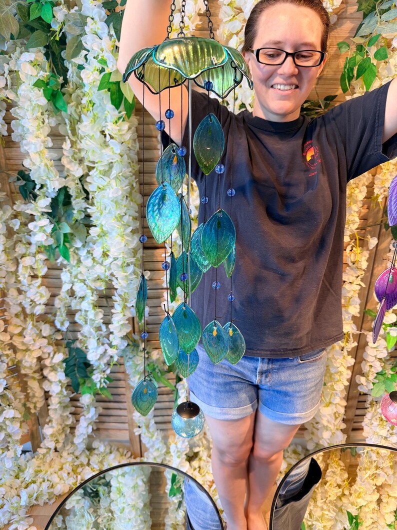 A woman is smiling and holding a colorful glass wind chime with green and blue leaves hanging from it.