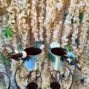 May include: Two bird-shaped metal planters with blue and gold accents. The planters have large eyes and beaks, and are perched on black metal stands. The background features white floral decorations.