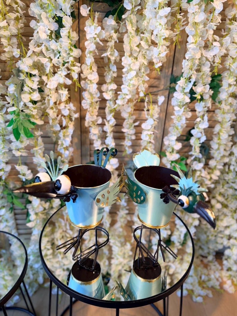May include: Two bird-shaped metal planters with blue and gold accents. The planters have large eyes and beaks, and are perched on black metal stands. The background features white floral decorations.