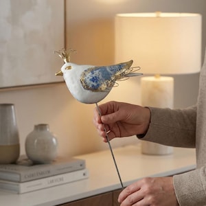 May include: A decorative metal bird figurine with a white body, blue and gold wing accents, and a gold crown. The bird is mounted on a thin metal rod, suitable for garden or home decor.