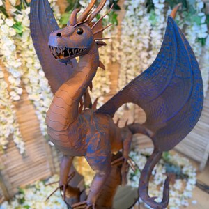 A large, detailed dragon sculpture with blue and orange hues stands prominently in the center of the image, surrounded by white flowers and greenery.