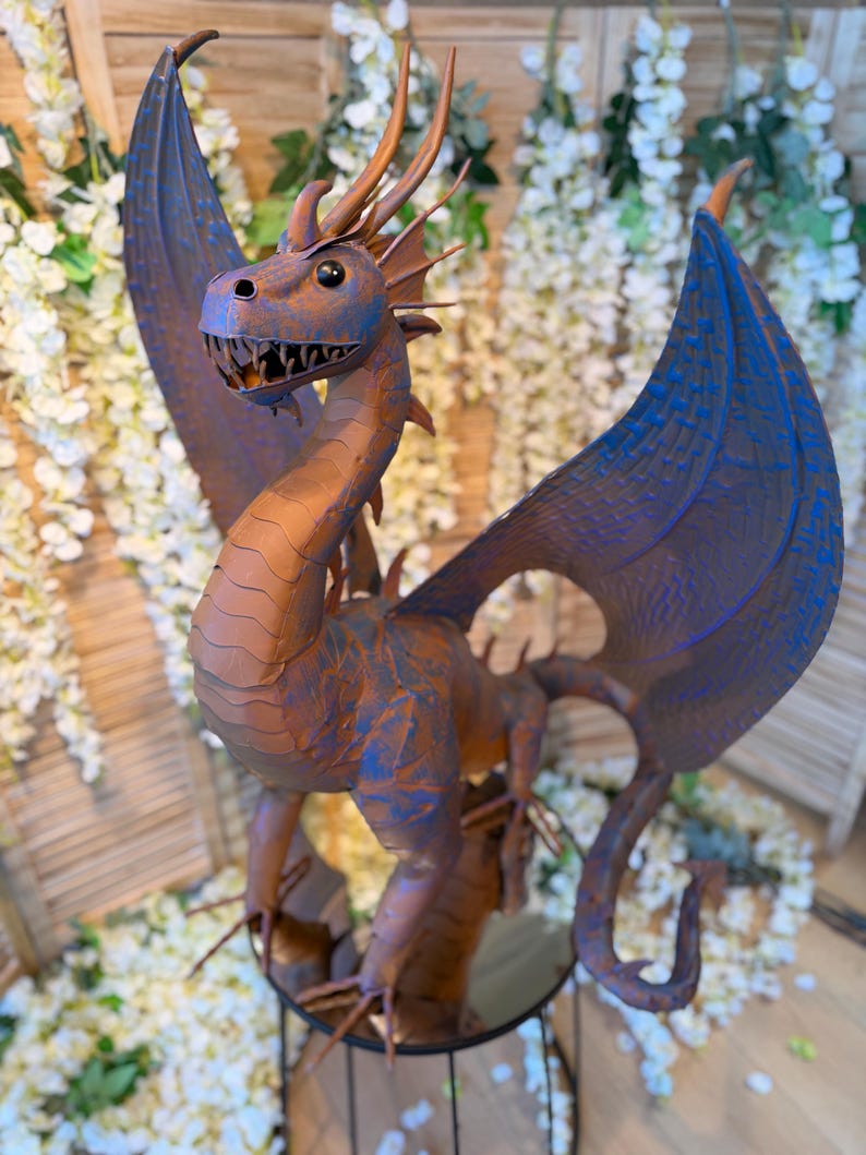 A large, detailed dragon sculpture with blue and orange hues stands prominently in the center of the image, surrounded by white flowers and greenery.