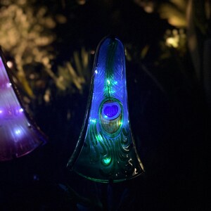 A tall, illuminated glass sculpture with a peacock feather design stands in a dark environment, surrounded by other illuminated objects.