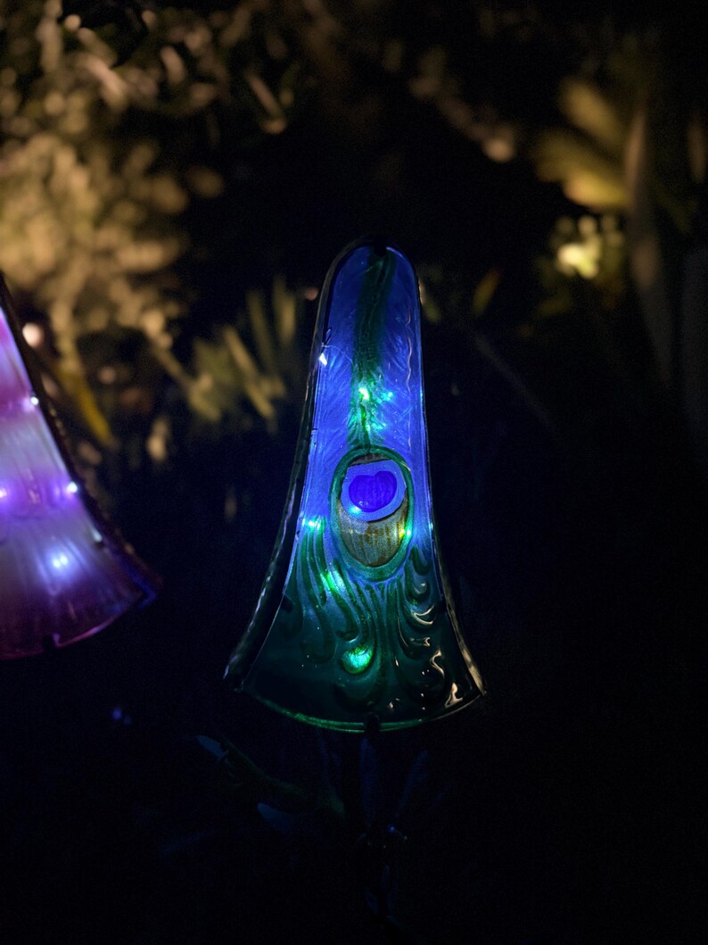A tall, illuminated glass sculpture with a peacock feather design stands in a dark environment, surrounded by other illuminated objects.