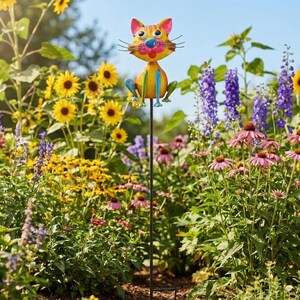A colorful garden cat figurine is perched on a metal pole amidst a vibrant garden filled with sunflowers, purple flowers, and other blooming plants.