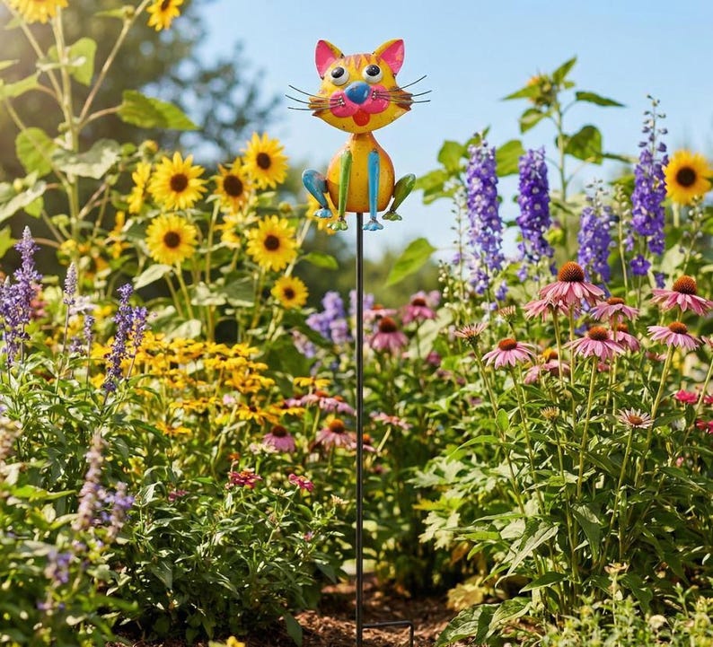 A colorful garden cat figurine is perched on a metal pole amidst a vibrant garden filled with sunflowers, purple flowers, and other blooming plants.