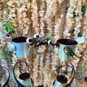 May include: Two whimsical, light blue metal bird planters with gold trim and decorative wings. The birds have large, googly eyes and are perched on black metal stands. The background features white flowers.