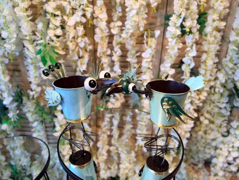 May include: Two whimsical, light blue metal bird planters with gold trim and decorative wings. The birds have large, googly eyes and are perched on black metal stands. The background features white flowers.